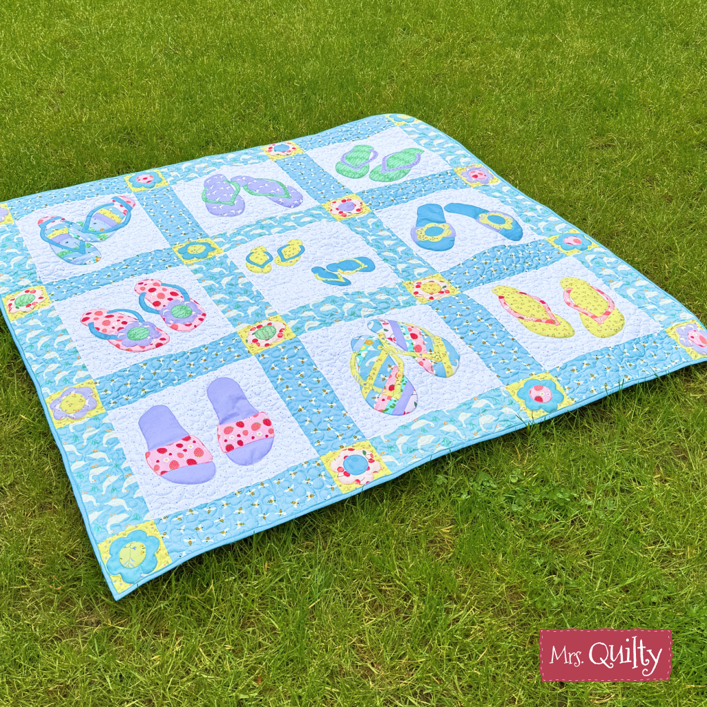 A playful blue and white Flip Flop Quilt, made from the downloadable PDF quilt pattern, is shown on green grass with creative quilting and a "Mrs. Quilty" label in the lower right corner.