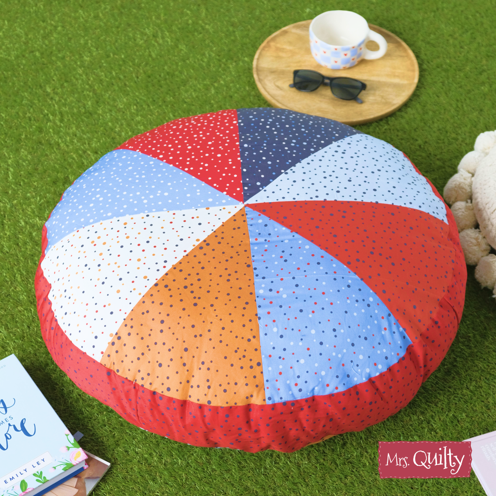 Quilted and round, this floor cushion features multicolored polka dots and rests on green grass. Nearby are a tray with a cup and sunglasses, plus books and a plush toy—an inviting look using the Floor Cushion Downloadable PDF Quilt Pattern.