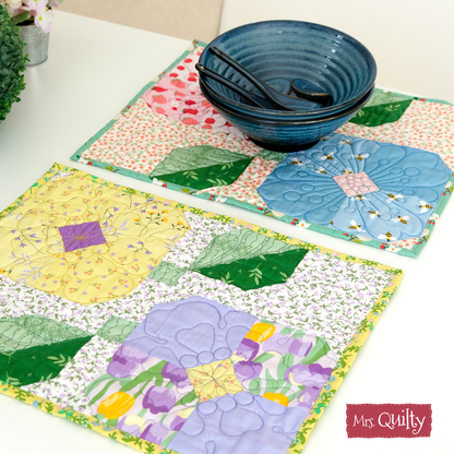Two colorful quilted placemats with floral designs, made from the "Flower Placemats Downloadable PDF Quilt Pattern," are shown with a blue ceramic bowl and black chopsticks on a white surface; "Mrs. Quilty" label appears in the corner.