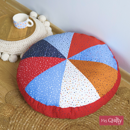 Floor cushion made with a colorful, dotted quilted pattern using the "Floor Cushion Downloadable PDF Quilt Pattern," shown on a wooden floor near a woven mat and polka dot cup. "Mrs. Quilty" logo in lower right. Confident beginner-friendly design.