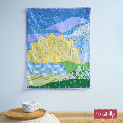 Brighten your space with the Sunrise Wall Hanging—a downloadable PDF quilt pattern featuring a sun, hills, clouds, and sky. Hang this charming "Mrs. Quilty"-labeled wall décor above a table for a cozy touch.
