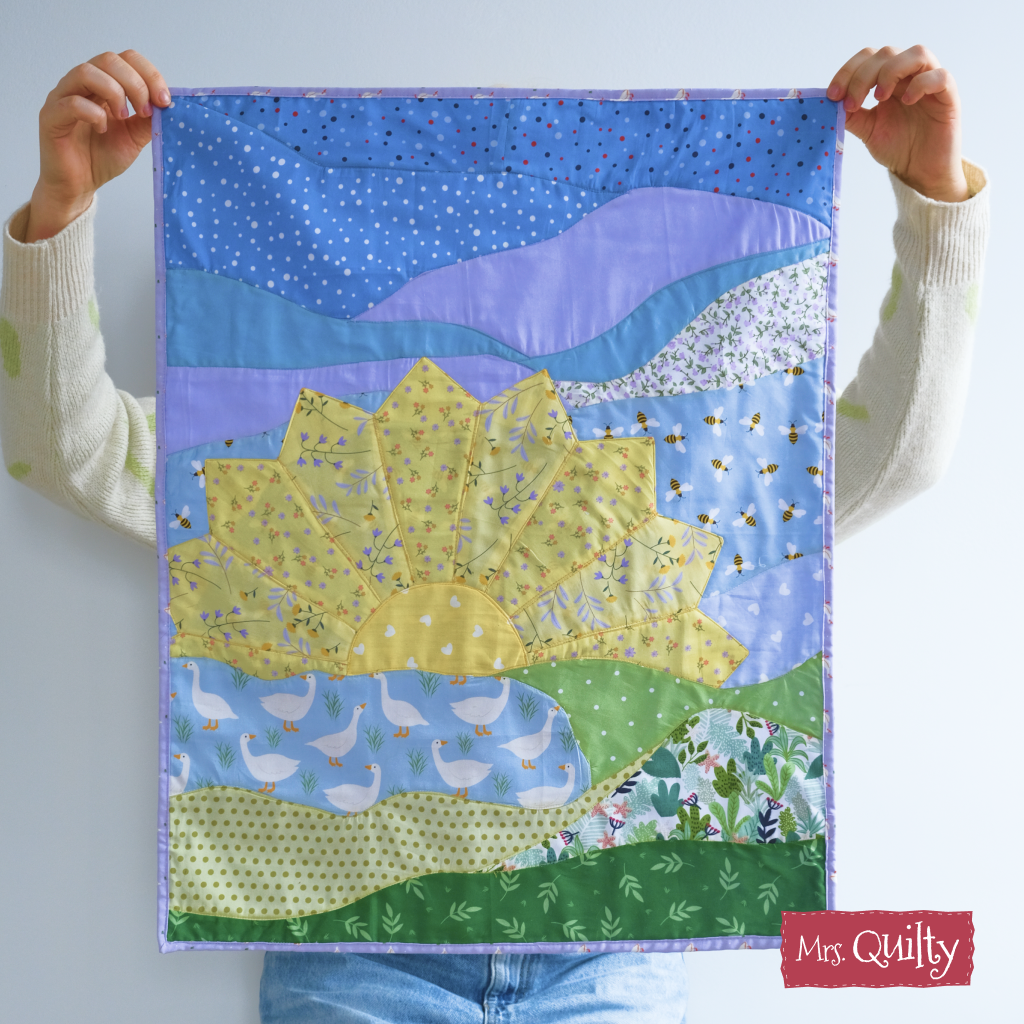 Person holding a handmade Sunrise Wall Hanging quilted with a large sunflower, blue sky, and patterned landscape; features "Mrs. Quilty" logo in the corner. Pattern available as a downloadable PDF quilt pattern.