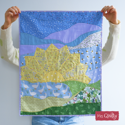 Person holding a handmade Sunrise Wall Hanging quilted with a large sunflower, blue sky, and patterned landscape; features "Mrs. Quilty" logo in the corner. Pattern available as a downloadable PDF quilt pattern.