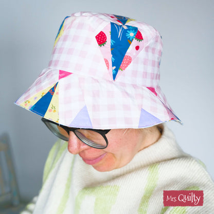 A person models a light pink checkered bucket hat featuring colorful patchwork triangles and a charming quilting pattern, created using the "Bucket Hat Downloadable PDF Quilt Pattern"; "Mrs. Quilty" logo is shown in the bottom right corner.