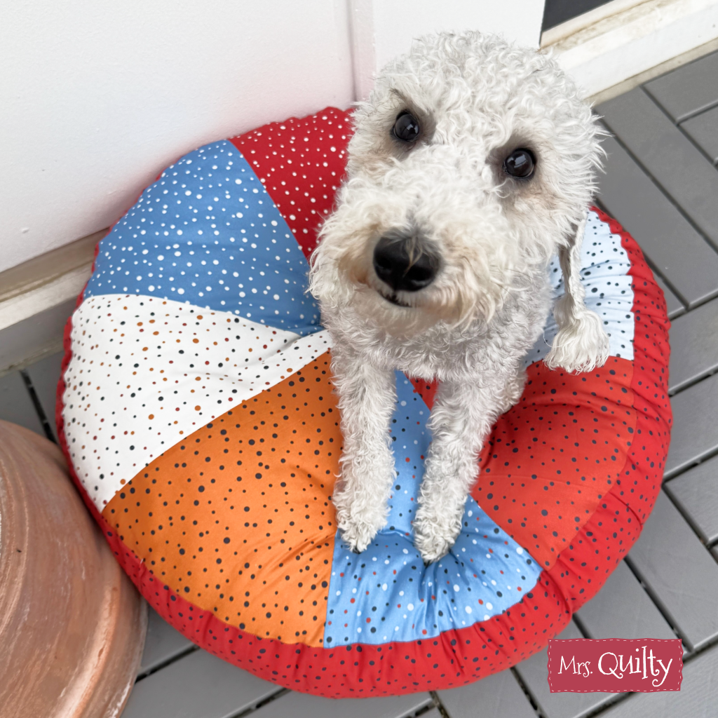 A small curly-haired dog sits on a round, colorful floor cushion made using the Floor Cushion Downloadable PDF Quilt Pattern, featuring customizable fabric options and suitable for confident beginner-friendly home decor.