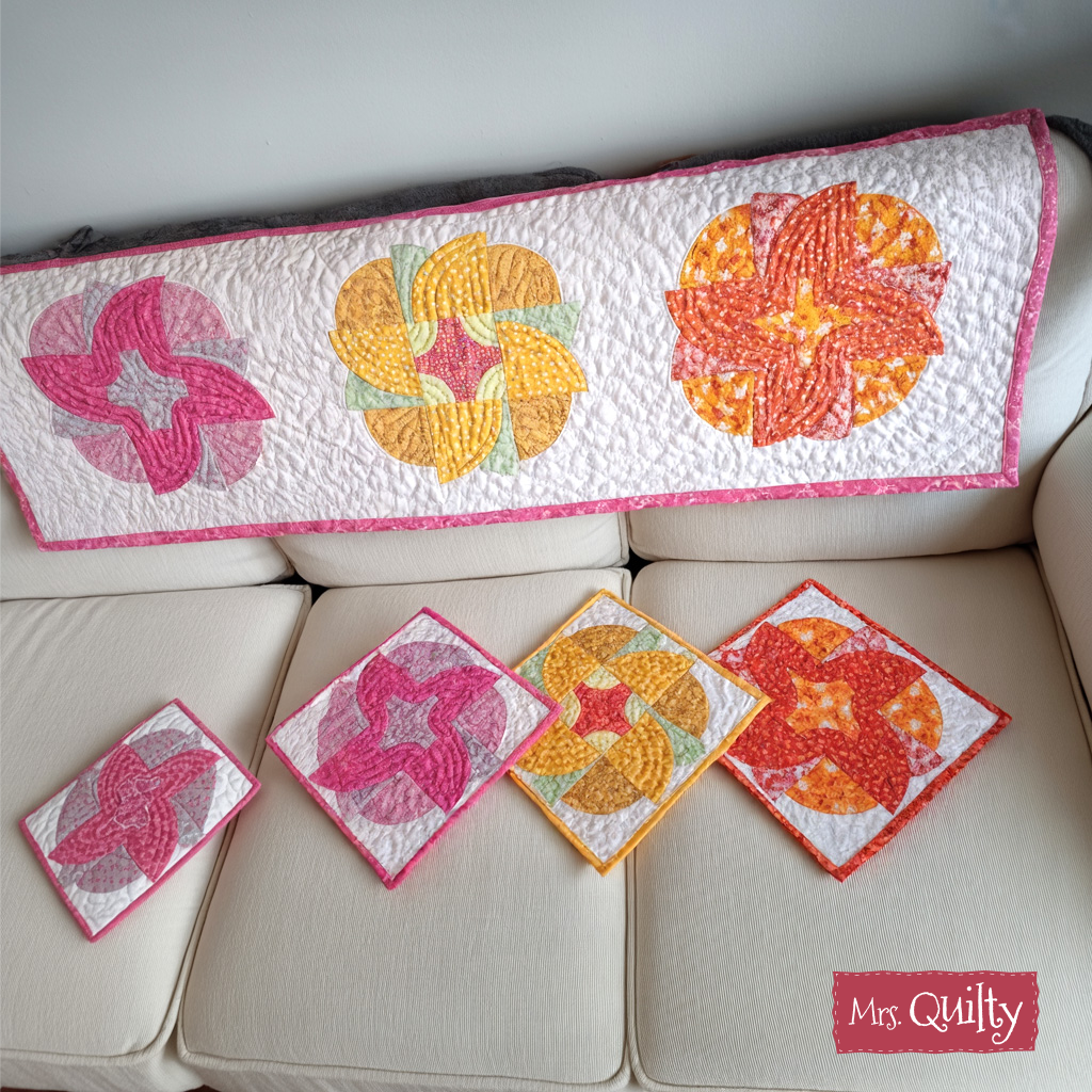 The "Blazing Blooms Table Runner and Mug & Rugs" PDF quilt pattern features a vibrant pinwheel table runner with floral appliqué and four matching coasters. Displayed on a sofa, it includes the text "Mrs. Quilty" in the lower right corner.