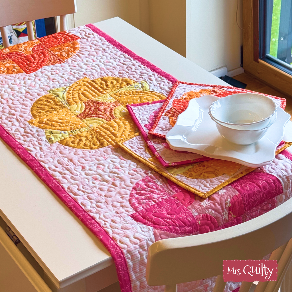 A white table features crisp dishes and quilted décor made with the Blazing Blooms Table Runner and Mug & Rugs Downloadable PDF Quilt Pattern in pink and orange.