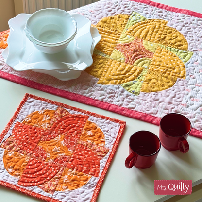The Blazing Blooms Table Runner and Mug & Rugs Quilt Pattern showcases orange and yellow floral appliqué designs, displayed with red cups and white dishes on a white table. Downloadable PDF pattern included.