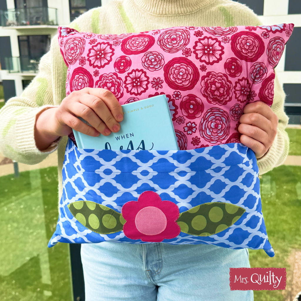 A person holds a Reading Pillow made with the Downloadable PDF Quilt Pattern, featuring floral prints, charming appliqué details, a handy book pocket, and the "Mrs. Quilty" logo.