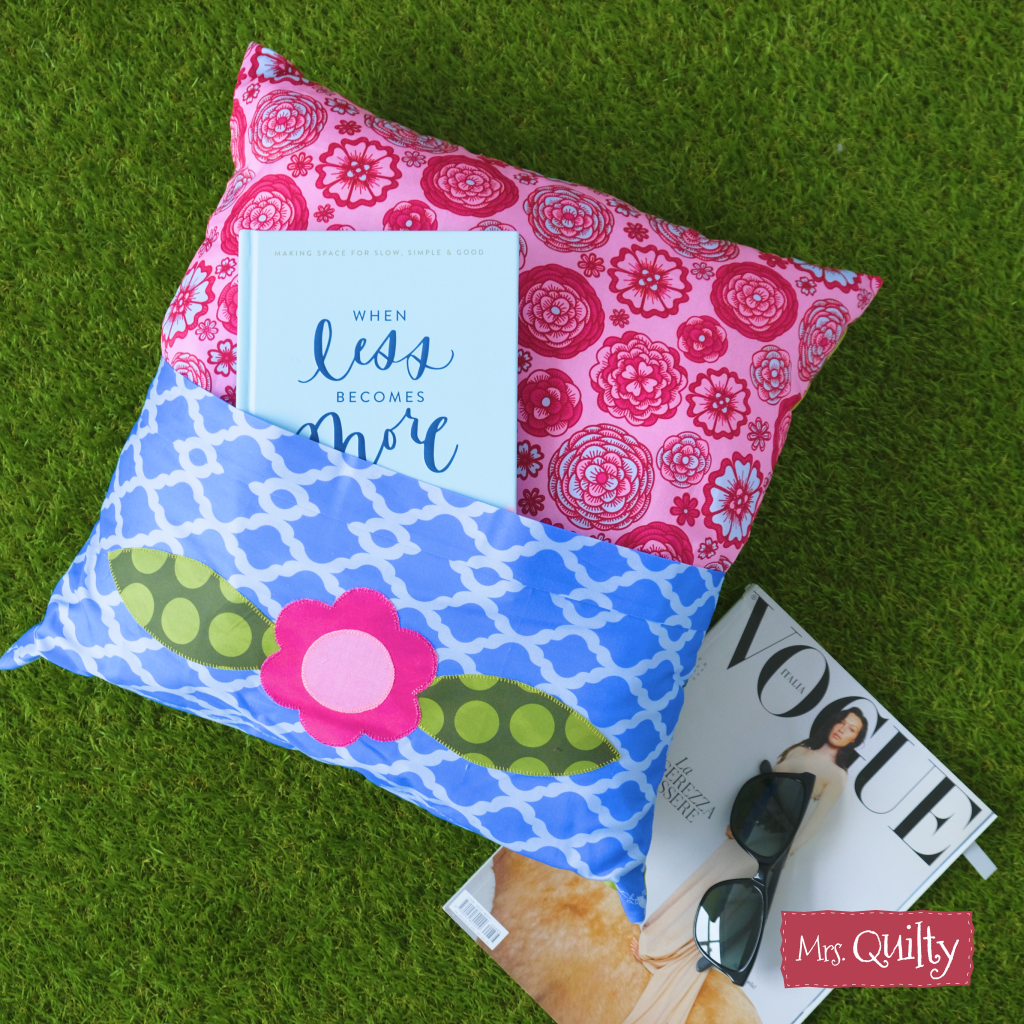 Two patterned pillows, a book, and sunglasses with Vogue magazine rest on the grass. Explore Mrs. Quilty – Summer Bundle of 7 Quilted Patterns, perfect for your next beginner-friendly quilts. The Mrs. Quilty logo is in the bottom right corner.