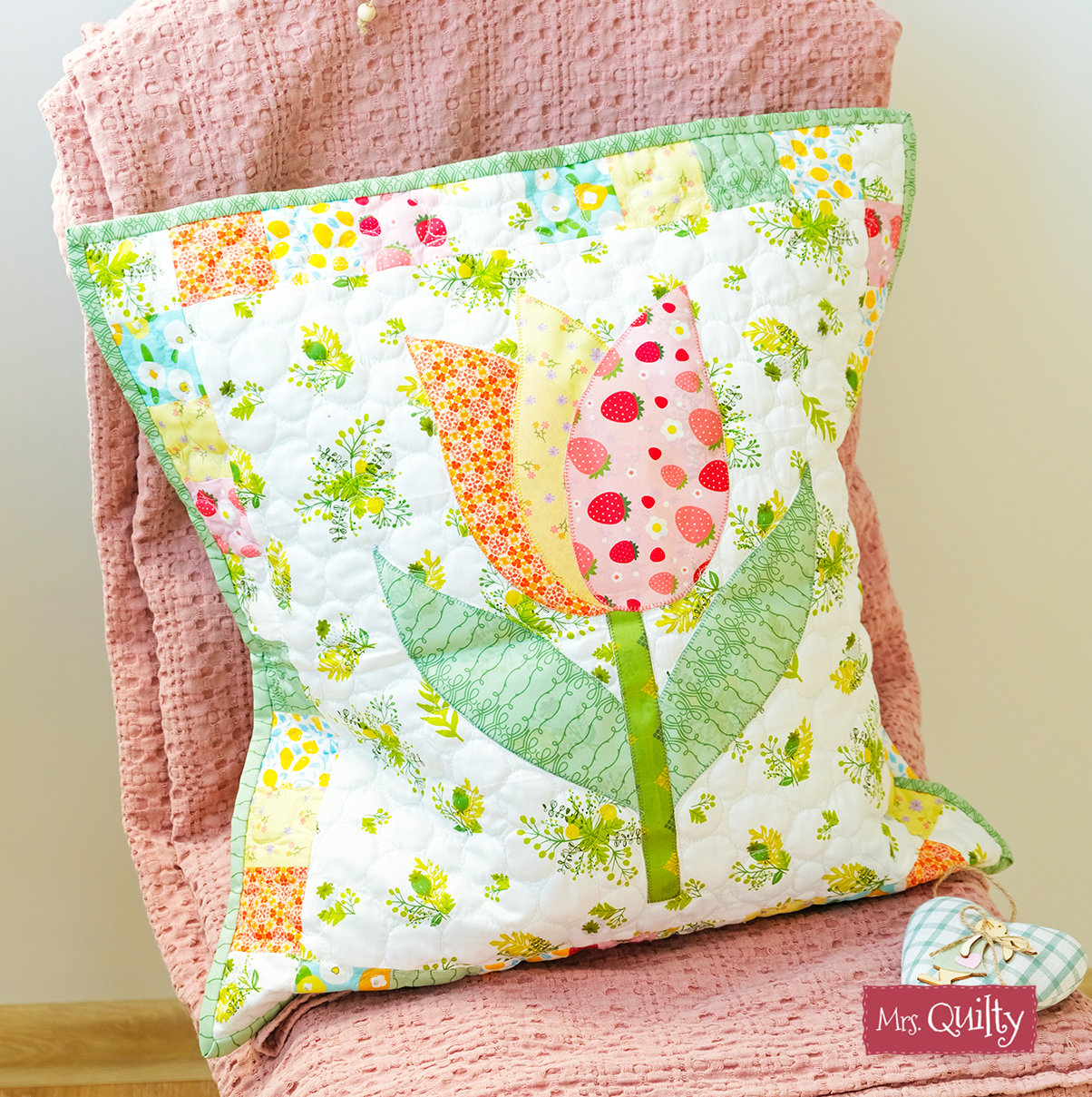 A decorative pillow with a floral quilted tulip design sits on a textured pink blanket. The "Mrs. Quilty" tag, perfect for seasonal decor, is visible at the bottom right. Inspired by Spring Magazine's 8 Quilting Patterns PDF download.