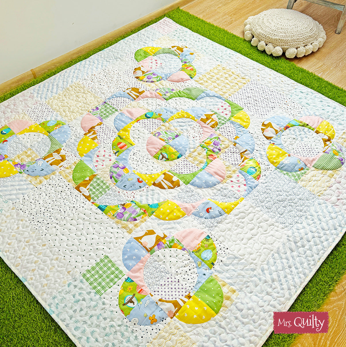A vibrant quilt with circular animal and geometric patterns adorns the wooden floor, accompanied by a green rug and round cushion, enhancing festive home decor. Discover more with our Spring Magazine – 8 Quilting Patterns Inside! Downloadable PDF.