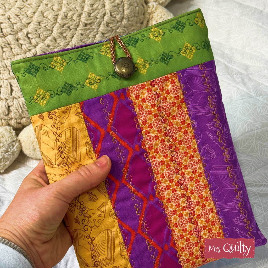 A hand holds a colorful quilted book sleeve made with the Book Sleeve Downloadable PDF Quilt Pattern, featuring green, purple, orange, and gold designs, a button and chain closure, and a small "Mrs. Quilty" label.
