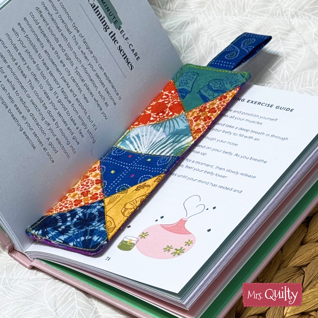 A Bookmark Downloadable PDF Quilt Pattern rests on an open book with illustrations and text, showing the “Mrs. Quilty” logo in the corner. Ideal for personal projects or as a thoughtful gift for any quilting enthusiast.