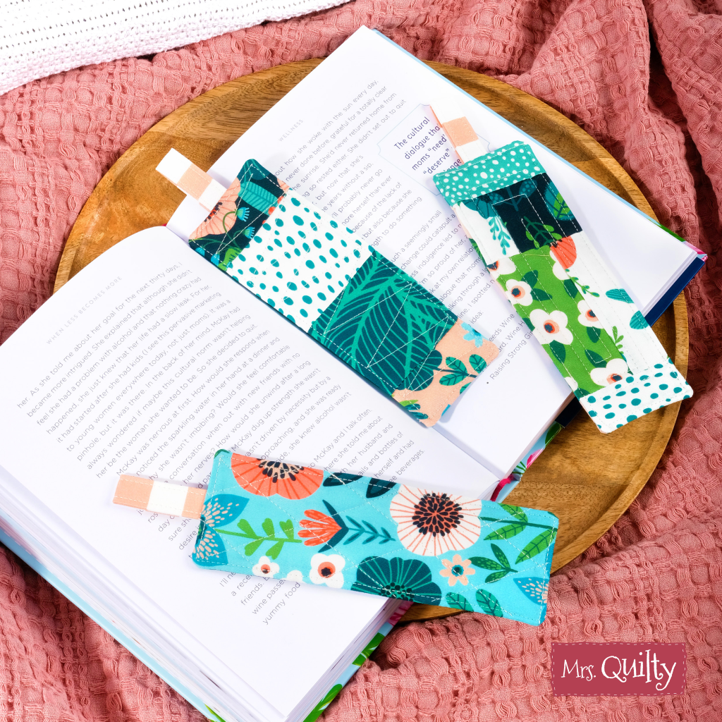 Three downloadable PDF quilt pattern bookmarks with colorful, floral designs rest on an open book atop a textured pink surface, featuring a "Mrs. Quilty" label—perfect personalized gifts for any book lover.