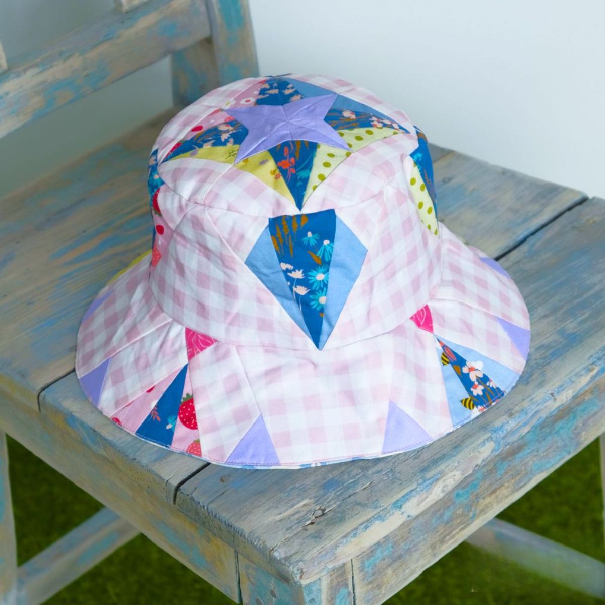 A downloadable PDF quilt pattern for a colorful patchwork bucket hat with geometric designs is displayed on a weathered blue wooden chair.