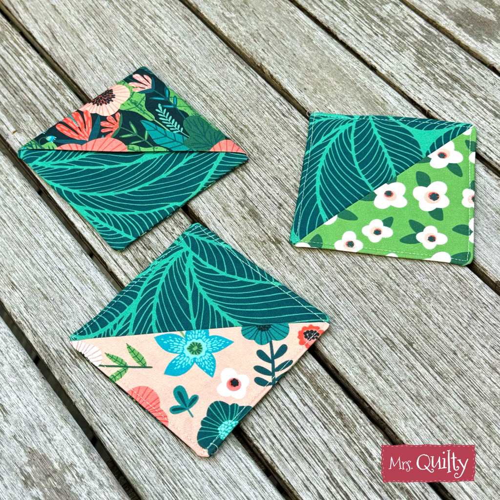 A set of three fabric coasters featuring floral and leaf patterns from quilting scraps is arranged on a rustic wooden surface. Each coaster showcases two contrasting designs. Product: Corner Bookmark Downloadable PDF Quilt Pattern.