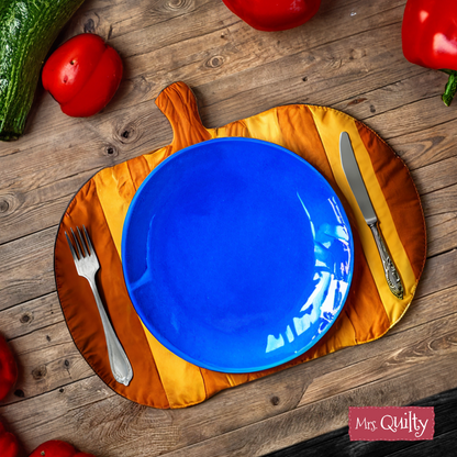 A blue plate, fork, and knife sit on a Halloween Pumpkin Placemat Quilt Pattern (downloadable PDF)—a festive fall decor—on a wooden table with red peppers and zucchini nearby.