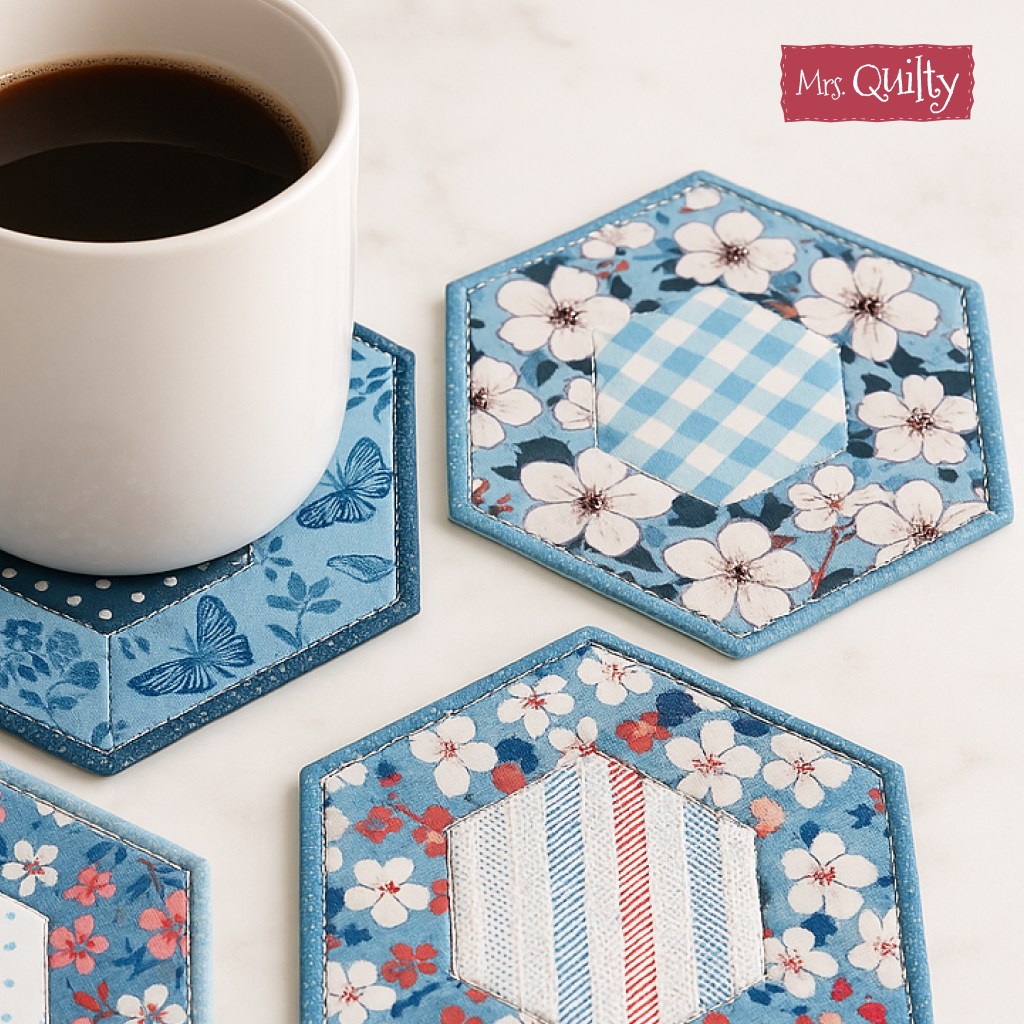 A white coffee mug rests on a blue hexagonal coaster featuring a butterfly motif, made with the Kitchen Coaster Downloadable PDF Quilt Pattern, beside two similar coasters with blue floral and geometric prints.