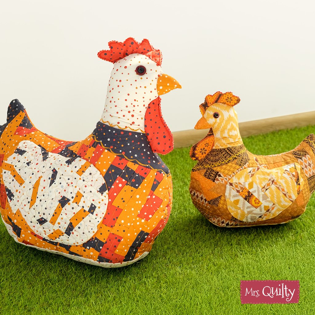 Two fabric chickens with colorful patchwork sit on artificial grass, adding farmhouse charm to any space. The "Mrs. Cluckworth Downloadable PDF Quilt Pattern" logo appears in the bottom right corner.