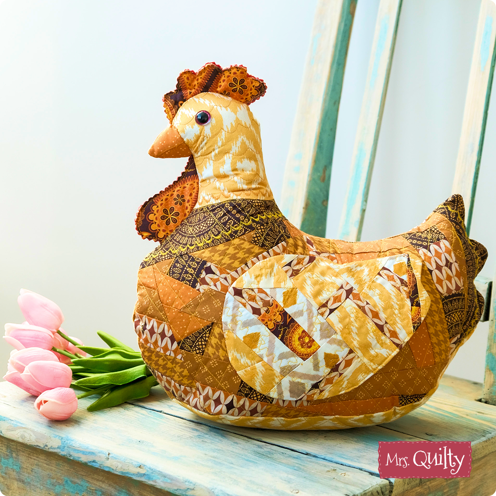A patchwork fabric chicken sits on a blue wooden chair beside pink tulips, bringing farmhouse charm; “Mrs. Cluckworth Downloadable PDF Quilt Pattern” is noted in the corner.