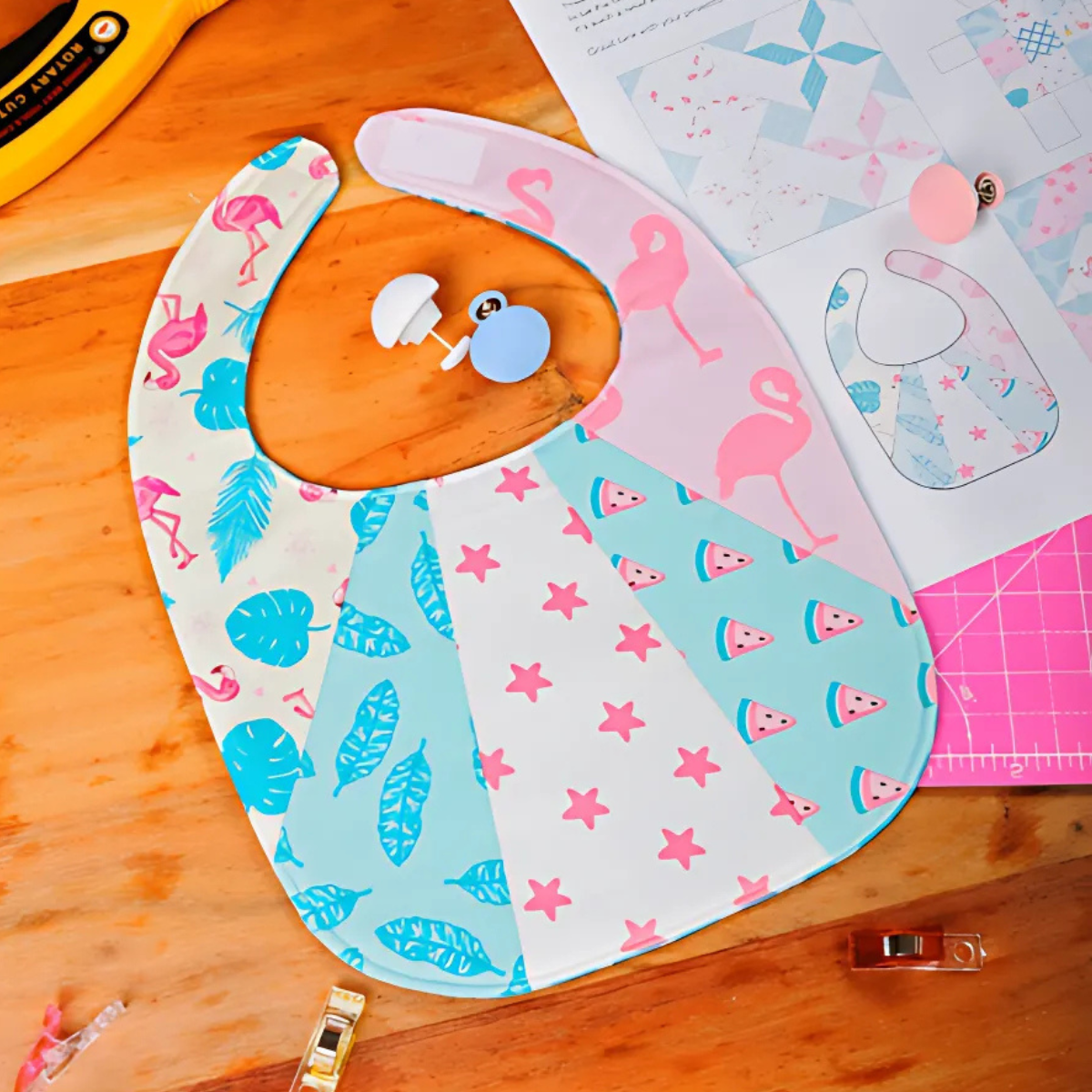 A brightly patterned patchwork baby bib featuring flamingo, star, leaf, and watermelon designs sits with sewing supplies and the Patchwork Baby Bib Downloadable PDF Quilt Pattern on a wooden table.