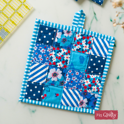 A charming kitchen accessory, the Pot Holder Downloadable PDF Quilt Pattern features blue and white patchwork with floral and geometric designs, displayed on a white surface beside a yellow ruler.