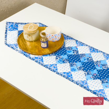 A wooden tray with a candle, jar, and white mug sits on a blue table runner made from the Table Runner Downloadable PDF Quilt Pattern atop a white table.