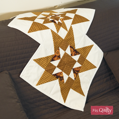 A Table Runner Downloadable PDF Quilt Pattern with brown star designs on white is shown on a dark brown sofa. The "Mrs. Quilty" label accents the corner. Ideal for home decor, this pattern is available as an instant PDF download.