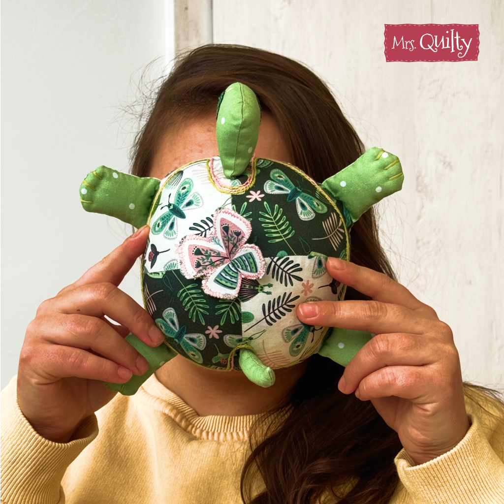 A person holds a plush quilted turtle toy in front of their face, obscuring their features. The toy is made using the "Timmy the Turtle Downloadable PDF Quilt Pattern," making it a charming choice for handmade gifts.