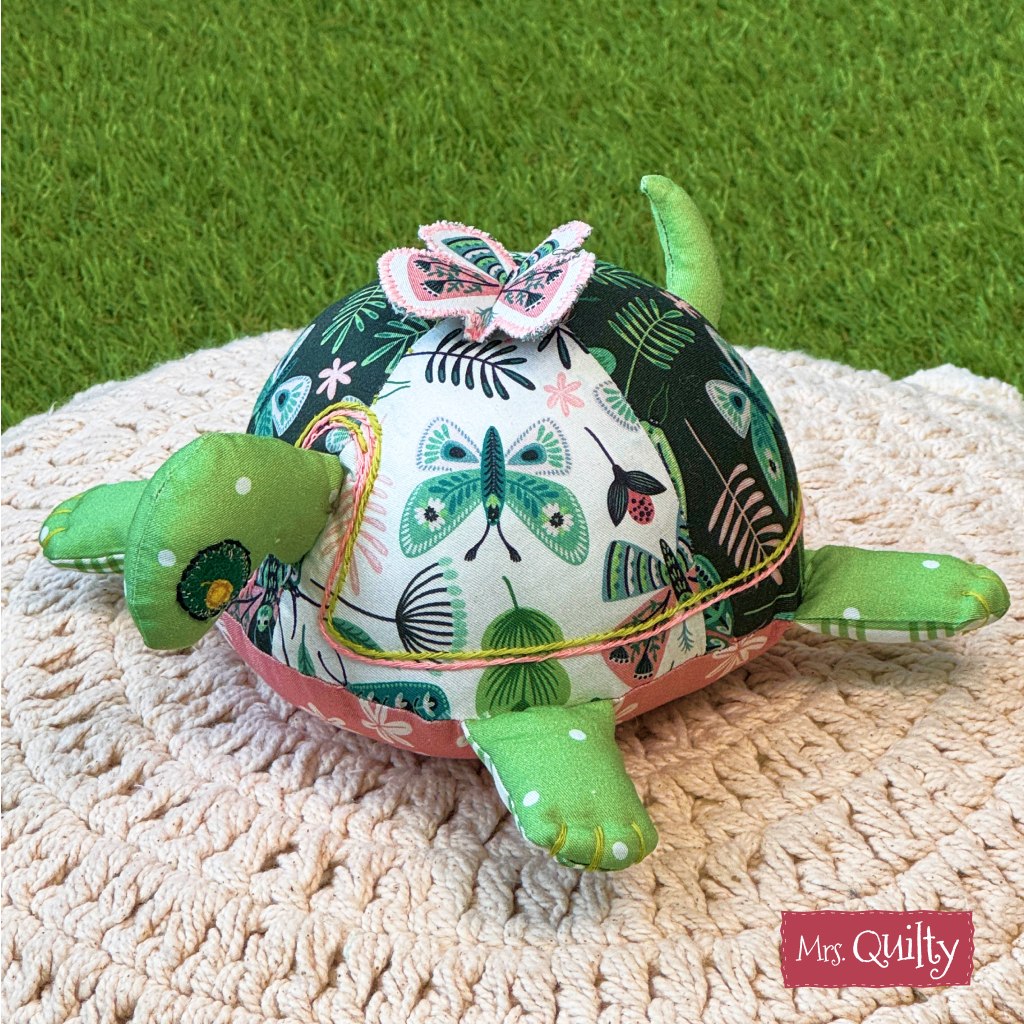 Timmy the Turtle Downloadable PDF Quilt Pattern features a quilted turtle with green flippers and a butterfly-print shell, perfect for beginners or handmade gifts. "Mrs. Quilty" appears in the corner, making it ideal for your next project.