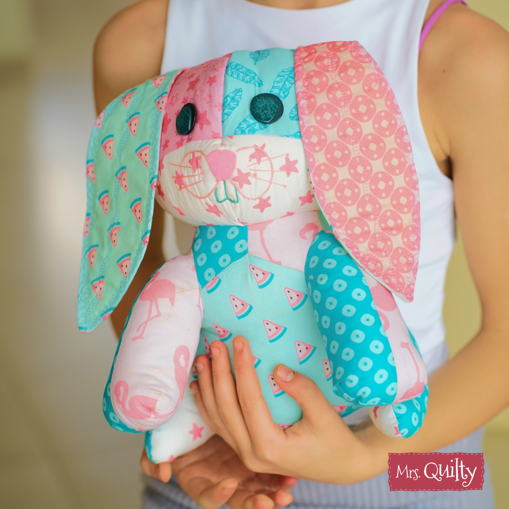 A person holds a stuffed bunny made with the “Bonnie the Bunny Downloadable PDF Quilt Pattern,” showcasing pink and blue patchwork, button eyes, and embroidery—a cute and beginner-friendly handmade gift idea.